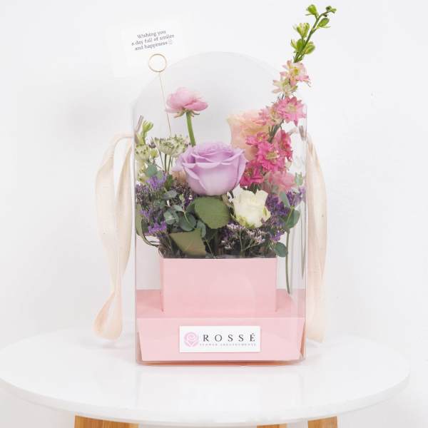 Pastel pink and purple floral arrangement in a pink box with clear cover and small greeting card.