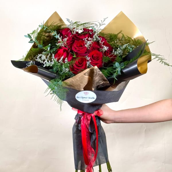 Hand-tied bouquet of red roses with white filler flowers wrapped in black and gold paper and red ribbon