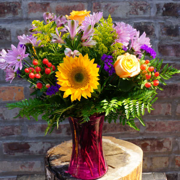 Mixed bouquet with a sunflower, roses, and purple daisies in a pink vase