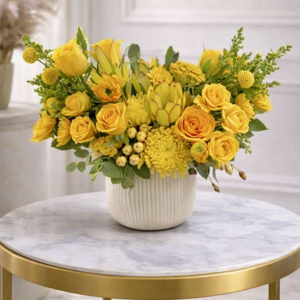 Low yellow flower arrangement with roses, protea, and mums in a white ceramic pot on a marble table