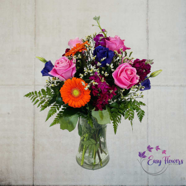 Mixed bouquet of pink roses, orange gerbera daisies, and blue blooms in a glass vase