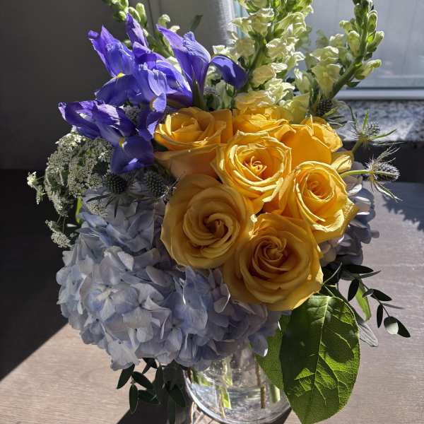 Bouquet of yellow roses, blue irises, and pale hydrangeas in a glass vase