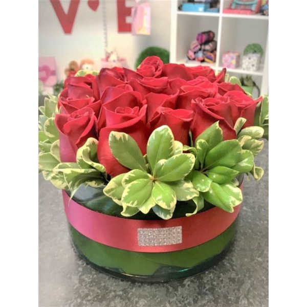 Low round arrangement of red roses with variegated green foliage in a glass vase wrapped with a pink ribbon
