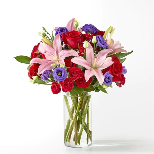 Mixed bouquet of pink lilies, red roses, and purple lisianthus in a clear glass vase.