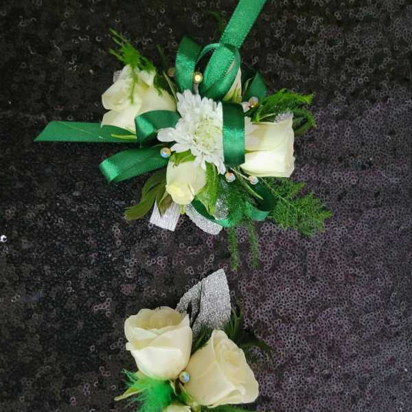 Two white rose corsages with green ribbon accents on a dark sequined background