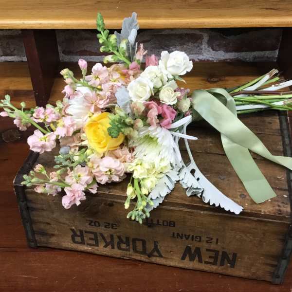 Loose hand-tied wedding bouquet of pastel blooms with a yellow rose and sage ribbon on a wooden crate.