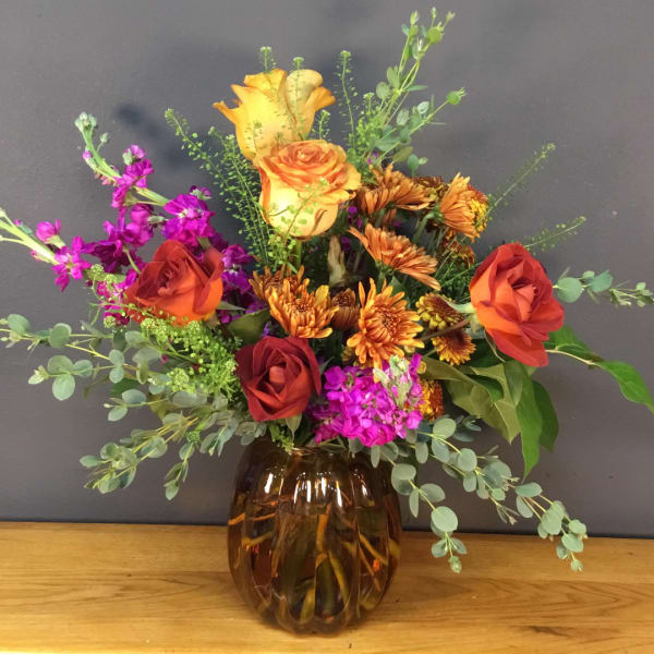 Mixed bouquet of orange and red roses with purple blooms in an amber vase.