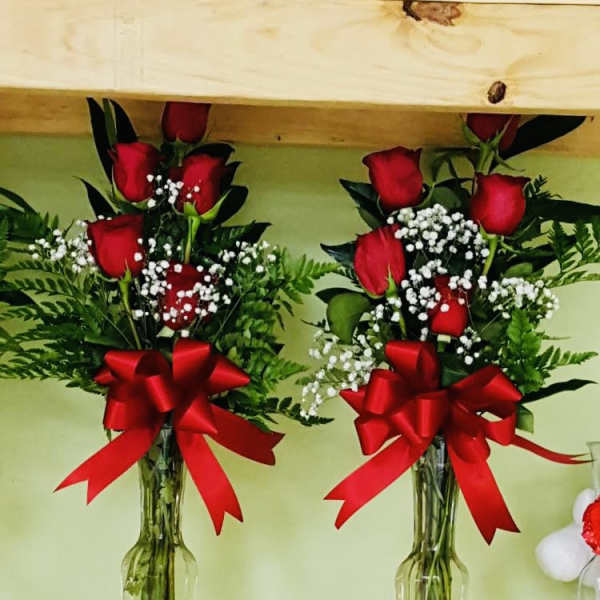 Two tall vases of red roses with white filler flowers and red bows on a wooden shelf