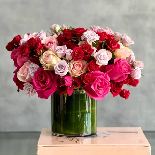 Low glass vase filled with dense mix of pink, red, and pastel roses on a light box.