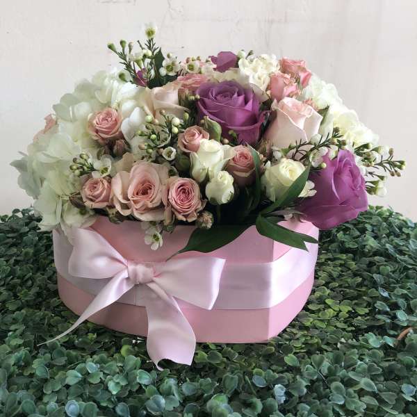 Round pink hatbox of white, blush, and lavender roses with a satin bow on a green textured surface