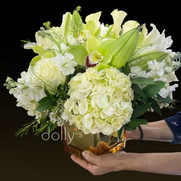 Low arrangement of white and pale green hydrangeas, roses, lilies, and an orchid in an amber glass cube vase.