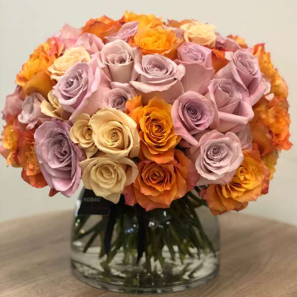 Compact arrangement of orange, lavender, and cream roses in a round clear glass vase on a wooden surface