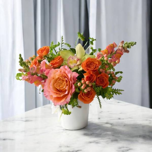 Compact arrangement of orange roses, pink blooms, and a green orchid in a white vase