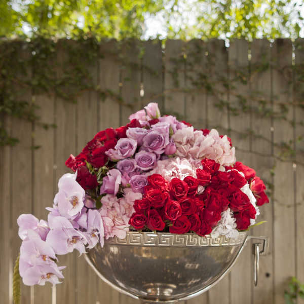 Pink, red, and lavender roses arranged in a silver urn with orchids draping over the side