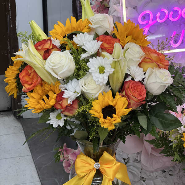 Bouquet of sunflowers, roses, lilies, and white daisies in a glass vase with a yellow bow