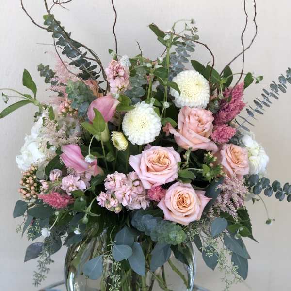 Tall pastel arrangement of pink roses, white mums, and calla lilies in a round glass vase