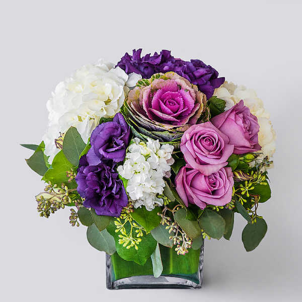 Compact arrangement of purple and lavender roses, lisianthus, and white hydrangeas in a square glass vase