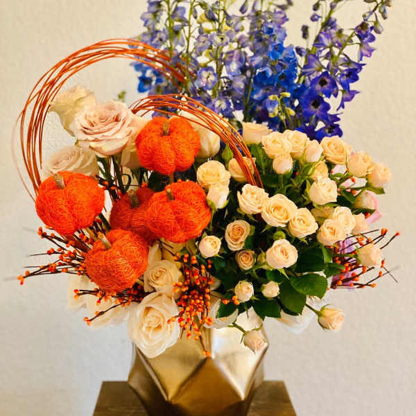 Bouquet of cream roses and blue delphinium with orange pumpkin accents in a gold vase