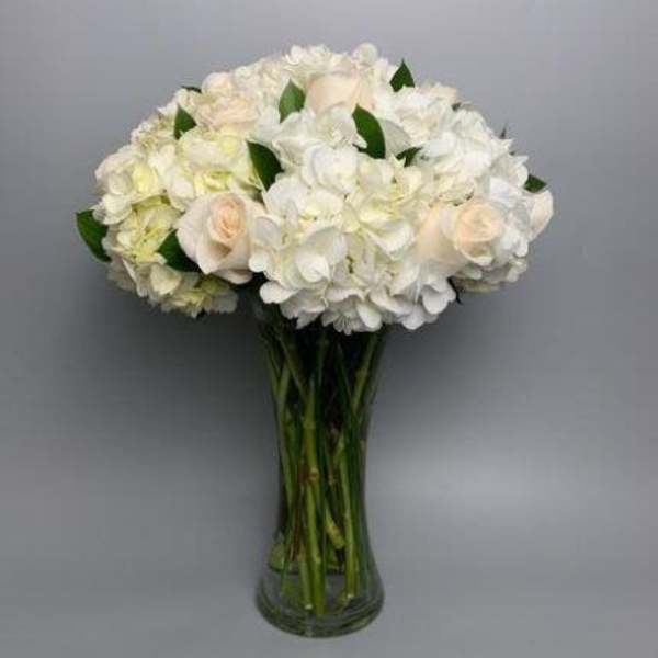 Tall clear glass vase filled with white hydrangeas and ivory roses in a rounded arrangement