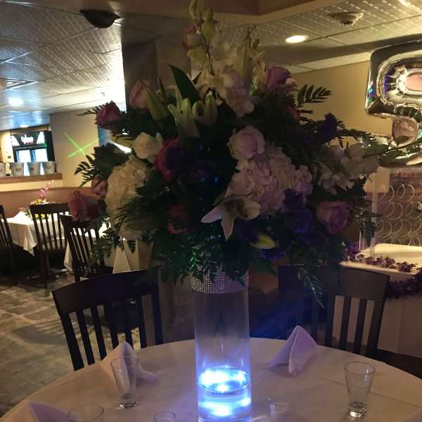 Tall floral centerpiece with pink, white, and purple blooms in a lit glass vase