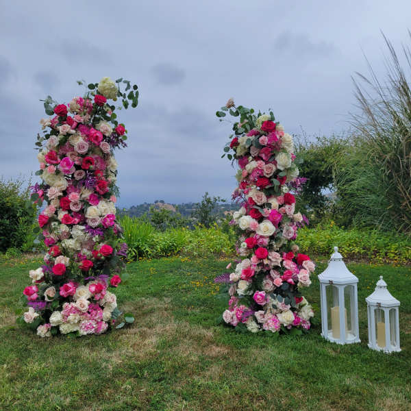 Two tall outdoor floral columns of pink and white roses with white lanterns on the grass