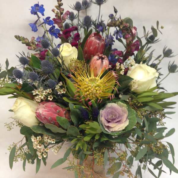 Mixed bouquet with protea, roses, and blue thistle in a burlap-wrapped vase