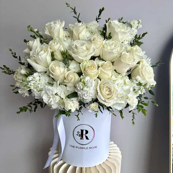 Round hatbox arrangement of ivory roses and white flowers with a ribbon in a white container