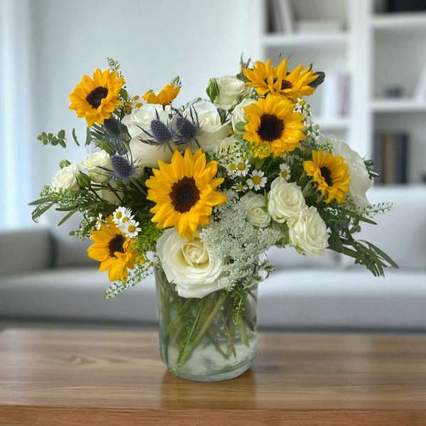 Yellow sunflowers and white roses arranged with small daisies in a clear glass vase on a wooden table