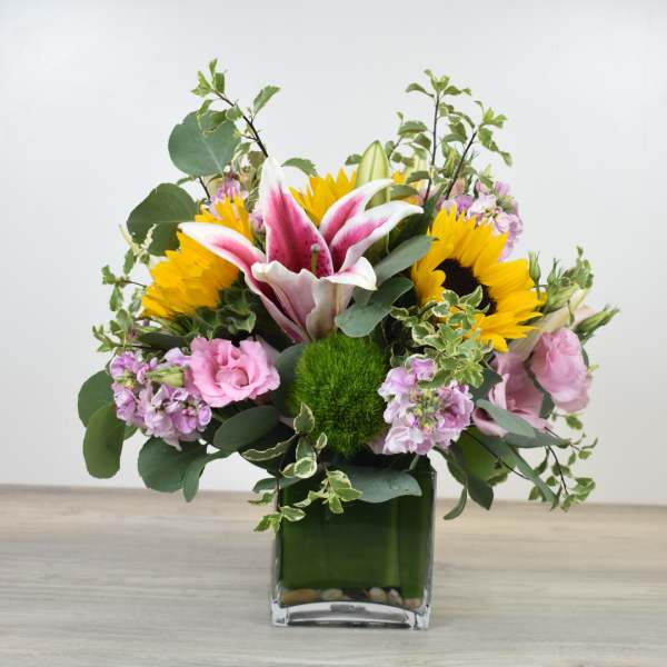 Bright mixed arrangement with sunflowers, pink lilies, and pastel blooms in a square glass vase