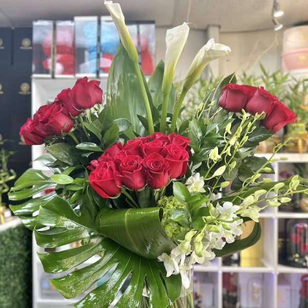 Tall arrangement of red roses, white calla lilies, and orchids in a clear glass vase with large glossy leaves