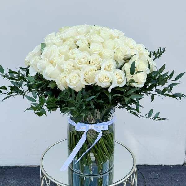 Large arrangement of cream roses with a collar of dark foliage in a tall clear cylinder vase.