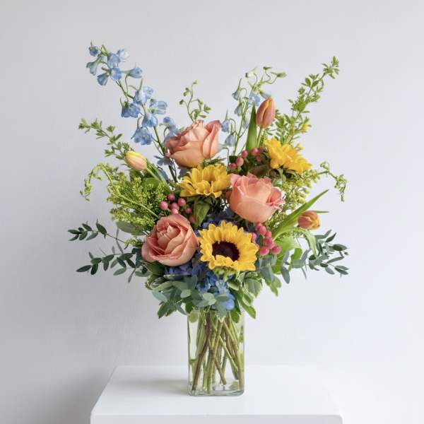 Tall mixed bouquet with yellow sunflowers, peach roses, blue blooms and tulips in a clear glass vase.