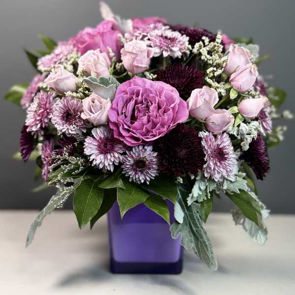 Rounded arrangement of pink roses and purple mums in a purple cube vase