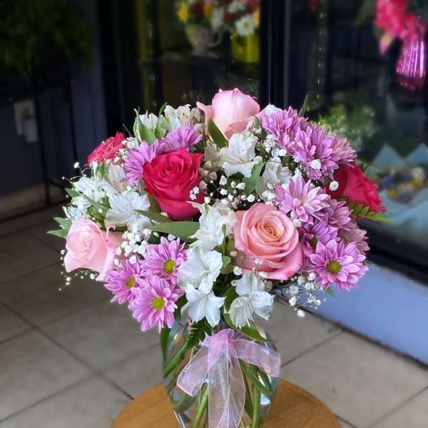 Pink roses, lavender daisies, and white blooms in a clear glass vase with a pink ribbon.