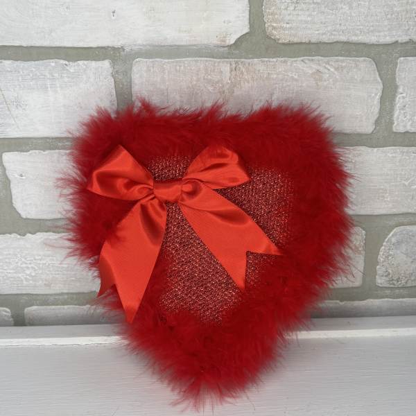 Red heart-shaped decorative pillow with feather trim and satin bow on a light brick background