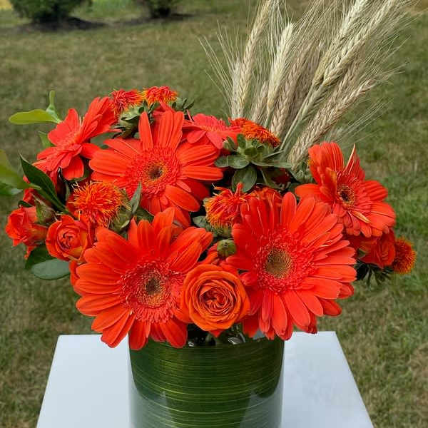 Orange gerbera daisies and roses in a glass vase with wheat stems