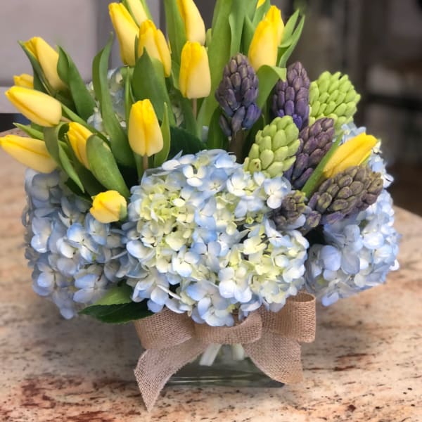 Yellow tulips and blue hydrangeas in a glass vase with a burlap bow