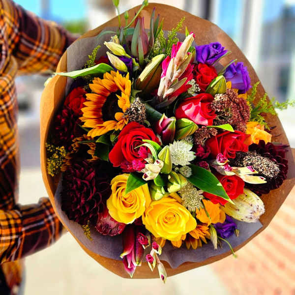 Handheld bouquet of mixed roses, sunflowers, and lilies in brown wrap