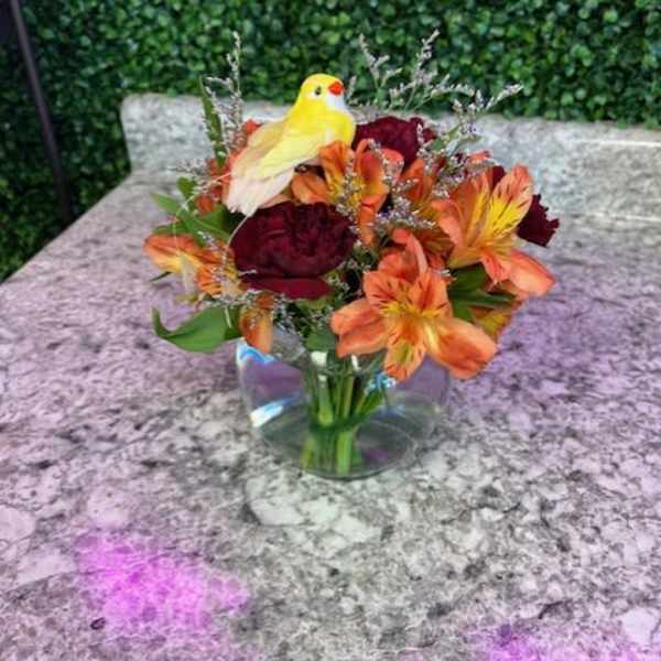 Bouquet of orange and burgundy flowers in a clear glass vase with a yellow bird figurine