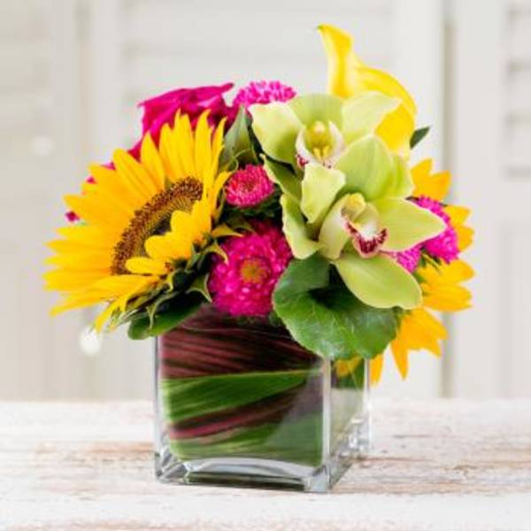 Bright mixed bouquet in a clear square vase with sunflowers and orchids