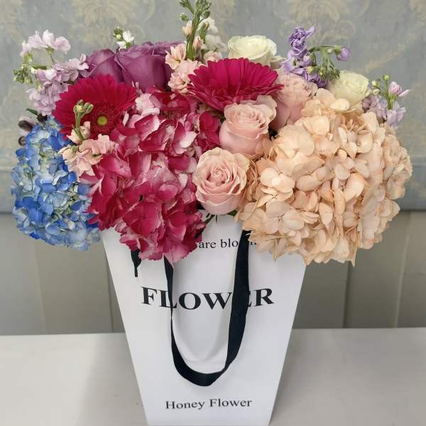 Mixed bouquet of hydrangeas, roses, and gerbera daisies in a white gift bag