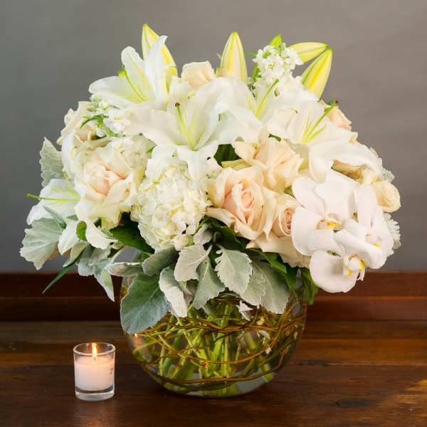 White lilies and pale roses in a glass bowl vase with a candle nearby