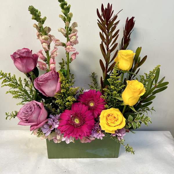 Rectangular arrangement of lavender roses, bright pink gerbera daisies, and yellow roses in a green container