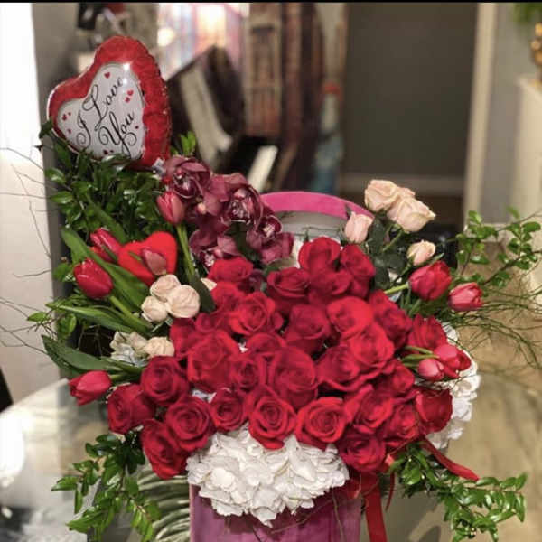 Round arrangement of red roses, tulips, orchids and white hydrangeas in a pink box with a heart balloon