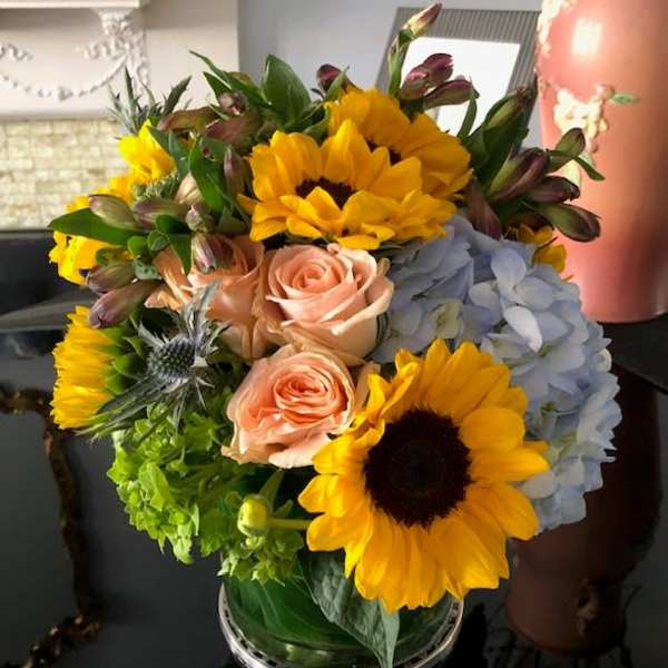 Bouquet of sunflowers, pale roses, blue hydrangea, and green blooms in a glass vase