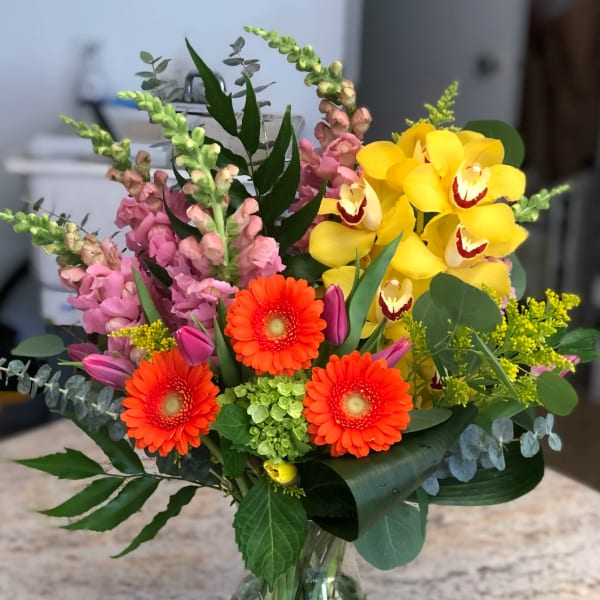 Mixed bouquet with yellow orchids, orange gerbera daisies, and pink snapdragons in a glass vase