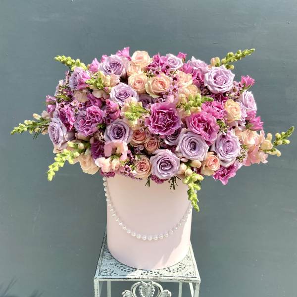 Lavender and pink rose arrangement in a pink hat box with pearls on a decorative metal stand.