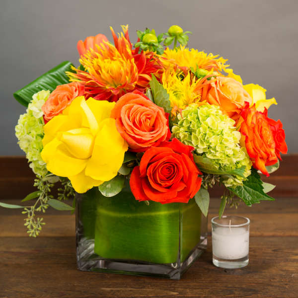 Bright orange, yellow, and red flowers in a square glass vase with a candle beside it.