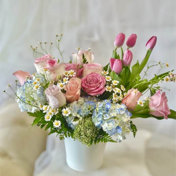 Pink roses and tulips arranged with blue hydrangeas in a white vase.