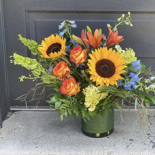 Mixed bouquet with sunflowers, roses, lilies, and blue flowers in a green vase